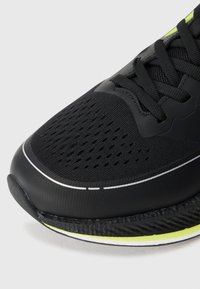 Shoe desportivo preto vista frontal com ventilação em malha, atacadores pretos, uma faixa refletora branca e uma sola com detalhes em amarelo, sobre um fundo branco.