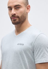 Lysegrå V-hals t-shirt lavet af blød bomuld, med en lille sort "GUESS"-logo på brystet. Korte ærmer og en afslappet pasform.