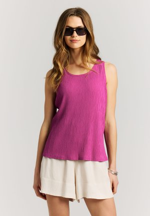 Woman with long wavy hair wearing black sunglasses, a textured sleeveless magenta top, beige shorts, and silver bracelets on her right wrist.