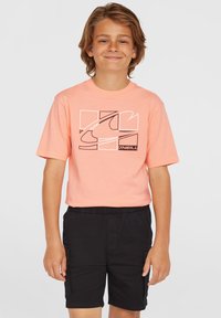 Ljusrosa t-shirt med svart grafisk design på bröstet, bärs med svarta cargoshorts. Tyget verkar mjukt och lätt.
