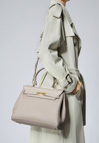 Sac à main en cuir beige avec une forme structurée, quincaillerie dorée, anse supérieure et bandoulière amovible. Porté avec un trench-coat clair.