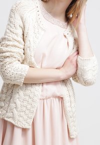 Krämfärgad chunky stickad cardigan med kabelmönster, öppen framsidan och ribbade ärmslut, lager på lager över en mjuk rosa klänning med en delikat halsringning.