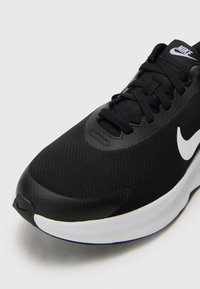 Chaussure de sport noire avec une tige texturée, logo swoosh blanc et une semelle blanche épaisse avec un bout arrondi. Les lacets sont noirs et plats.