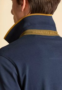 Polo di colore blu navy con colletto alto. Accenti di contrasto giallo e verde oliva. Testo "AERONAUTICA MILITARE" ricamato sul colletto.