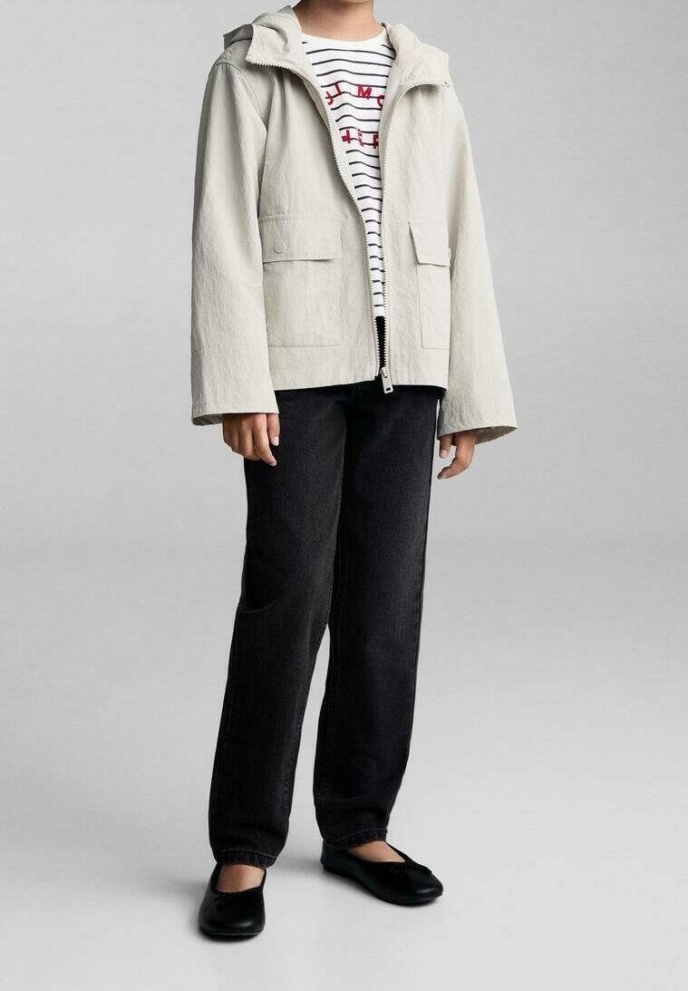 Personne portant une veste à capuche beige clair sur une chemise rayée, un pantalon noir et des ballerines noires, debout devant un fond uni.