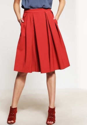 Mujer con falda plisada roja hasta la rodilla con bolsillos, combinada con sandalias rojas de tacón y punta abierta y una blusa azul metida dentro.
