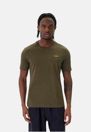 Uomo con capelli intrecciati che indossa una maglietta verde oliva con un piccolo emblema giallo alato sul petto sinistro e pantaloni scuri.