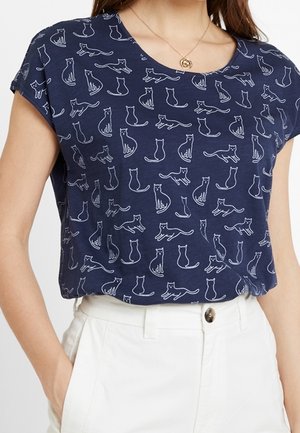 Granatowy top z krótkim rękawem ozdobiony białymi rysunkami przedstawiającymi różne pozy kotów, zestawiony z białymi spodniami z wysokim stanem i złotym naszyjnikiem z zawieszką.