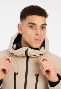 Beige waterdichte jas met een capuchon, zwarte ritsen, verstelbare trekkoorden en geperforeerde accenten. Het model houdt de trekkoorden vast.