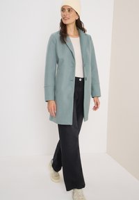 Ljusblå ullblandad kapp med avskuren krage, enknapps stängning och långa ärmar, matchad med svarta vida byxor och sneakers.