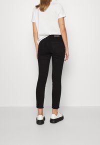 Jeans negros ajustados de cintura alta, con una textura suave, diseño de cinco bolsillos y un pequeño logo en la cintura. Combinados con zapatillas blancas.