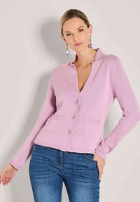 Frau in einem leicht lila Knopfpullover mit Taschen und blauen Jeans, die vor einem neutralen Hintergrund posiert.