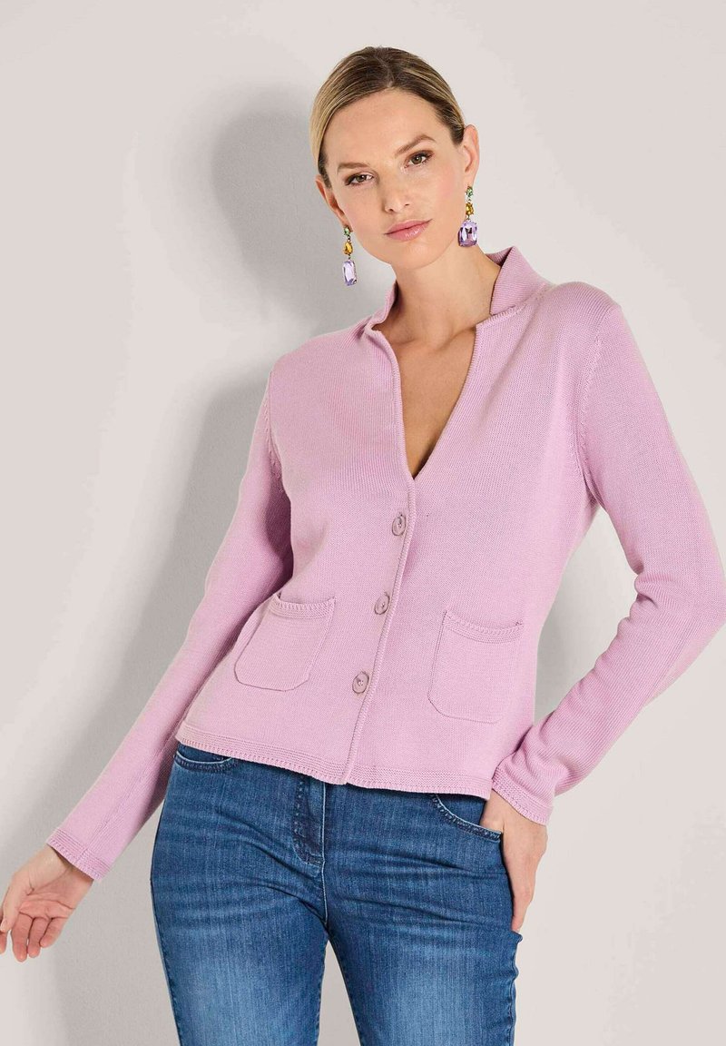 Frau in einem leicht lila Knopfpullover mit Taschen und blauen Jeans, die vor einem neutralen Hintergrund posiert.