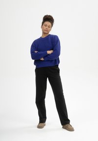 Blauer Strickpullover mit rundem Ausschnitt, kombiniert mit schwarzen Hosen. Braune Slipper. Das Modell posiert mit verschränkten Armen. Einfarbiger weißer Hintergrund.