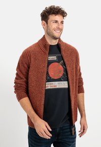Rustfarbener Strickcardigan mit Reißverschluss, geripptem Kragen und Bündchen. Getragen über einem schwarzen Grafik-T-Shirt. Texturiertes, lässiges Design.