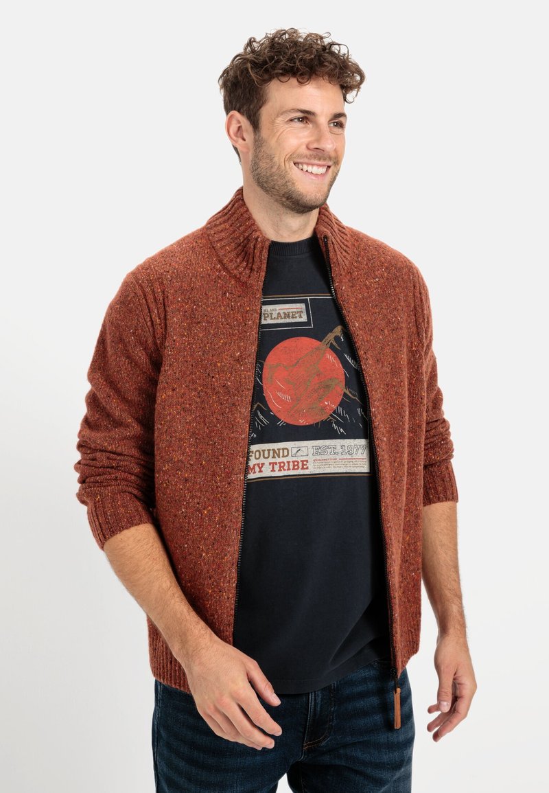 Roodkleurige gebreide cardigan met een rits aan de voorkant, ribbelkraag en manchetten. Draagt over een zwart graphic t-shirt. Gestructureerd, casual ontwerp.