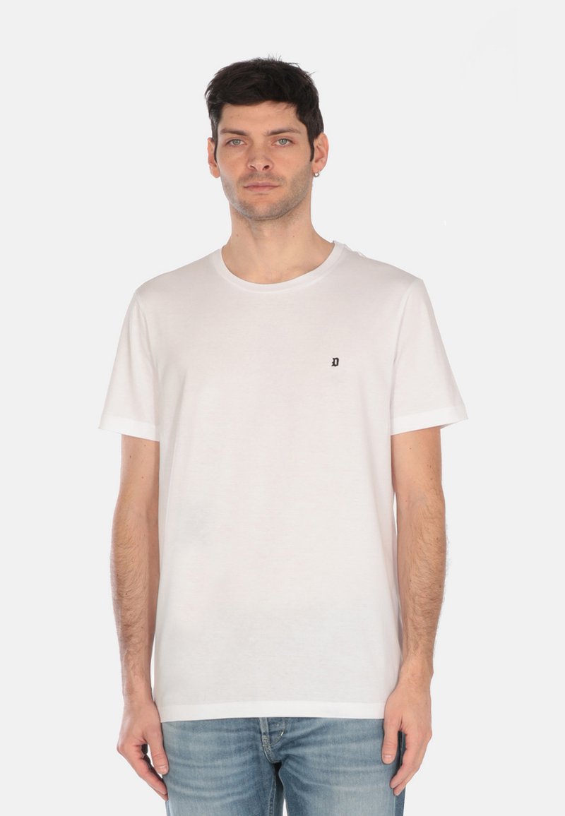 T-shirt bianca in cotone con scollo rotondo, maniche corte e un piccolo logo nero sul petto. Il modello indossa jeans blu.