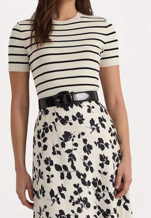 Frau trägt ein cremefarbenes Kurzarmhemd mit schwarzen horizontalen Streifen, einen schwarzen Gürtel und einen cremefarbenen Rock mit schwarzen Blumenmustern.