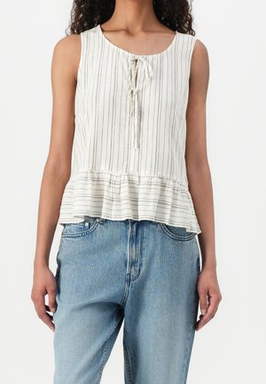 Kobieta ubrana w białą bluzkę bez rękawów z cienkimi pionowymi paskami, detalem wiązanym z przodu, falbankowanym dołem i jasnoniebieskimi dżinsami.