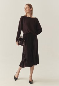 Donkerpaars gebreid truitje gecombineerd met een geplooide midi rok. Het model houdt een donkere leren clutch vast en draagt zwarte puntige hakken.