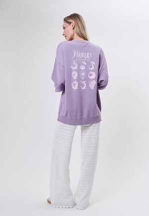 Kvinde i hvide bukser iført en lilla sweatshirt med teksten "Farmers Market" og frugtillustrationer på ryggen, stående mod en ensfarvet baggrund.