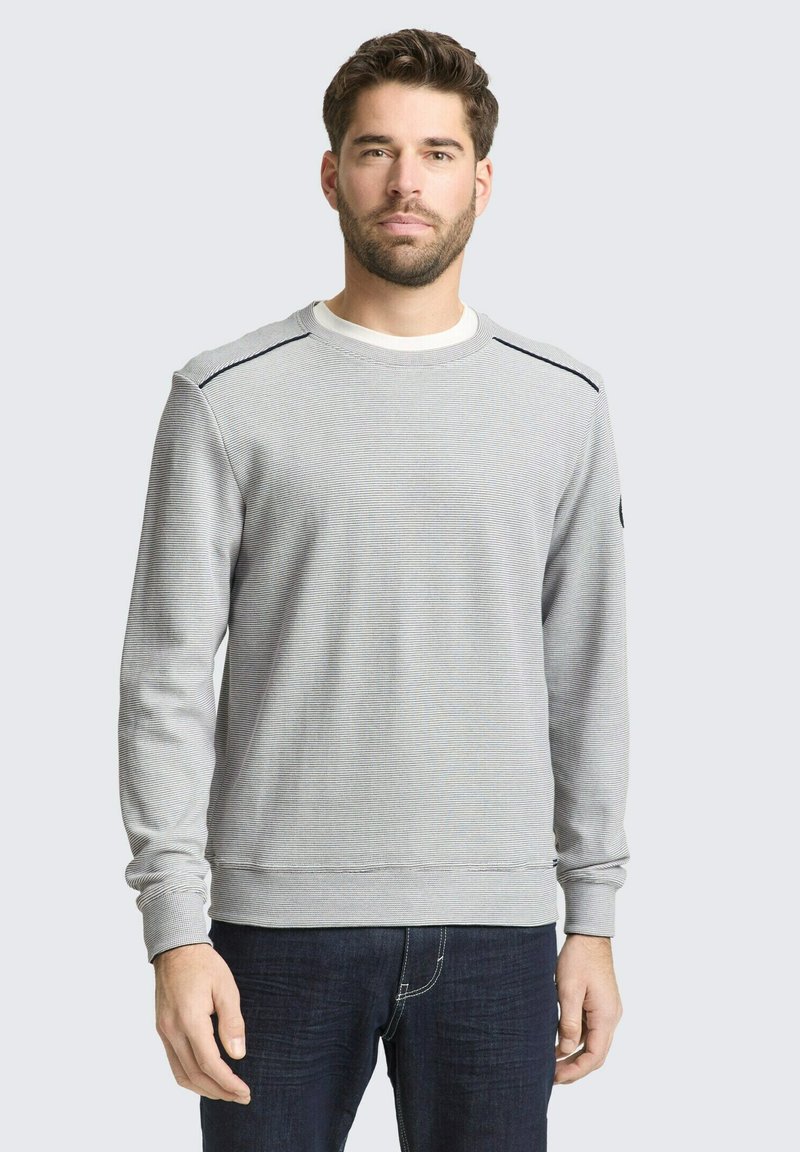 Muž s tmavými vlasmi a bradou, ktorý má na sebe svetlošedý rebrovaný sveter s dlhým rukávom a tmavomodré džínsy, stojaci pred jednoduchým pozadím.