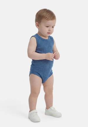 Tout-petit aux cheveux clairs debout, portant une combinaison sans manches bleue et des baskets blanches, regardant vers le bas avec les mains jointes.