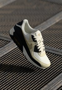 Weiße und schwarze Turnschuhe mit einer strukturierten Oberseite, abgerundeter Zehenpartie und sichtbarer Luftpolsterung, ergänzt durch graue Akzente und kontrastierende Schnürsenkel.