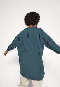 Übergroßes türkisfarbenes Sweatshirt mit langen Ärmeln, auf der Rückseite mit dunkelblauen Buchstaben „G R A W“ bedruckt und mit abgerundetem Saum. Weiche Textur.