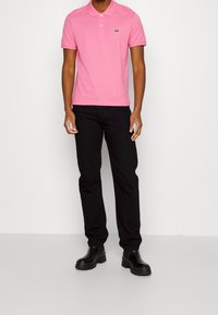 Polo en coton rose avec un logo vert sur la poitrine, associé à un jean noir et des bottines noires, posé devant un fond neutre.