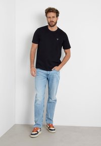 T-shirt nera in cotone con piccolo logo triangolare, abbinata a jeans in denim azzurro chiaro e sneakers multicolori con accenti arancioni.
