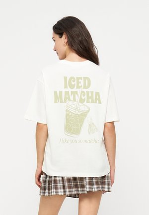 Kobieta o długich ciemnych włosach, ubrana w białą, oversize koszulkę z napisem „ICED MATCHA” i ilustracją szklanki, zestawioną z kratkowaną spódnicą.