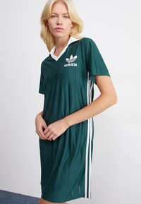 Zaļš sporta kleitas tērps ar baltām akcentiem, īsām piedurknēm, v-veida apkakli un vertikāli svītrotu tekstūru. Adidas logotips ir izvietots krūtīs.