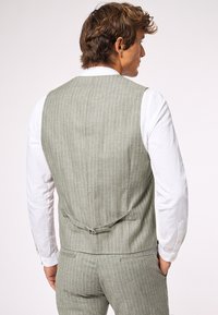 Helle grüne gestreifte Weste aus einem Leinen-Mischgewebe, mit einer Rückenschnalle und getragen über einem weißen Langarmhemd.