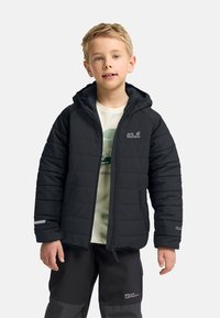 Chaqueta negra acolchada con capucha y logo en el pecho. Presenta bolsillos con cremallera y detalles grises en las mangas. Lleva puesta una camisa clara.