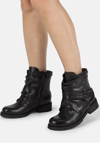 Botas de tobillo de cuero negro con un diseño arrugado, hebilla decorativa y correas texturizadas. Presentan un tacón bajo y cierre con cremallera.