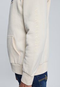 Sudadera beige con el bordado "valecuatro" a lo largo de la manga, con bolsillo tipo canguro y puños ribeteados. Material de mezcla de algodón.