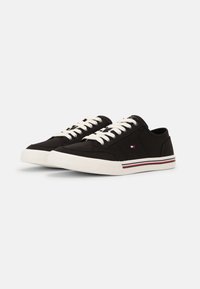 Schwarze Leinenschuhe mit weißen Schnürsenkeln, runder Zehenpartie und Gummisohle. Mit einem kleinen Logo und roten sowie blauen Streifenakzenten an der Sohle.