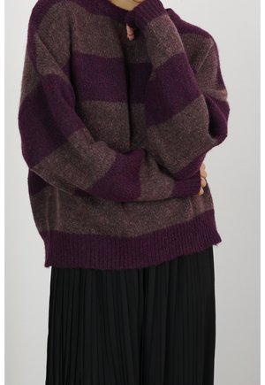 Henkilö pukeutuneena ylisuureen violettiin ja ruskeaan raidalliseen pörröiseen neuleeseen, jossa on pitkät hihat, ja mustaan lurelokynähameeseen, kädet ristissä kasvojen lähellä.