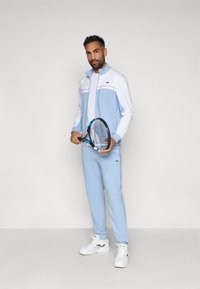 Ανοιχτό γαλάζιο τενιστικό κοστούμι με λευκές λεπτομέρειες και κόκκινες ραφές. Το μοντέλο κρατάει μία ρακέτα τένις και φοράει λευκά αθλητικά παπούτσια. Ορατό πράσινο λογότυπο.