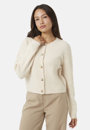 Femme aux longs cheveux foncés portant un cardigan en laine crème boutonné et un pantalon beige, posant avec une main près de sa clavicule.