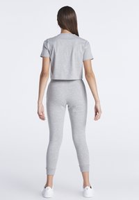 Mujer de pie de espaldas, vistiendo un top corto de manga corta gris, leggings de cintura alta a juego y zapatillas blancas sobre un fondo blanco.