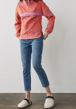 Fleece pullover in orange with pink leopard print, featuring a neck zipper and elastic cuffs. Paired with blue jeans and cream slip-on shoes.