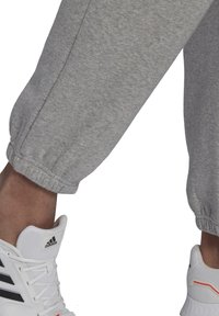 Šedé tepláky s elastickými manžetami, vyrobené z měkké tkaniny. V kombinaci s bílými teniskami Adidas s černými a oranžovými akcenty.
