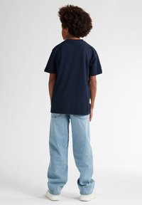 Niño con pelo rizado, vestido con una camiseta azul marino, jeans azul claro y zapatillas blancas, de pie de espaldas contra un fondo blanco liso.