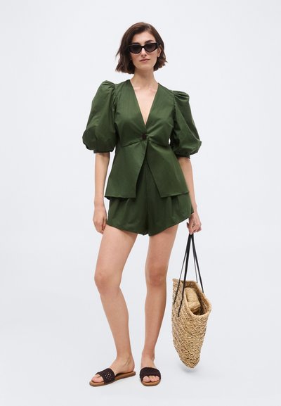 Γυναίκα που φοράει σκούρα πράσινη μπλούζα με φουσκωτά μανίκια και σορτς στο ίδιο χρώμα, μαύρα γυαλιά ηλίου, πλεκτά σανδάλια, κρατώντας μεγάλη πλεκτή τσάντα ώμου.