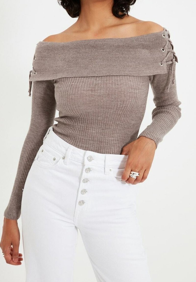 Femme portant un pull côtelé taupe épaules dénudées avec des détails lacés sur les côtés et un jean blanc taille haute à boutons, main posée sur la hanche.