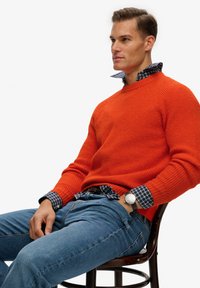 Suéter naranja con textura acanalada sobre una camisa de cuadros azul, combinado con vaqueros azules. La persona lleva un reloj negro en la muñeca.