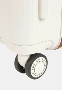 Valigia rigida bianca con un dettaglio in pelle marrone e ruote nere lisce e circolari con un logo bianco.