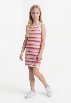 Niña joven con cabello rubio largo, vestida con un vestido de punto sin mangas a rayas y zapatillas blancas, de pie contra un fondo blanco.